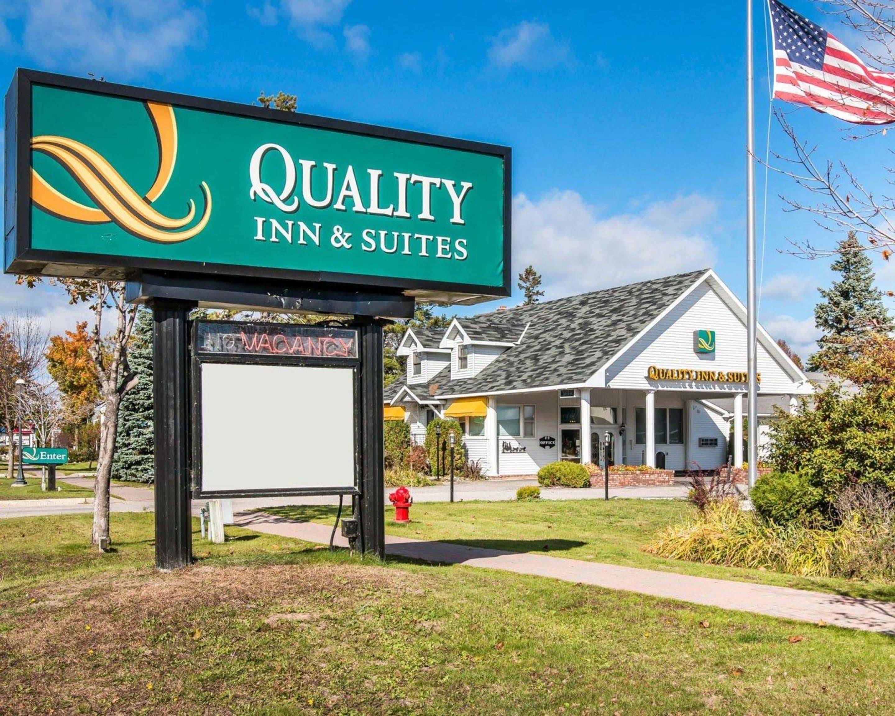 Quality Inn & Suites -hotellin kyltti, jossa on ilmoitus vapaasta huoneesta, punainen paloposti ja valkoinen rakennus, jossa on vihreä logo.