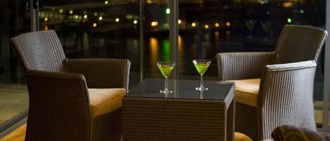 Uma área de estar com cadeiras de vime e uma mesa, dois copos de martini sobre a mesa, e vista para as luzes da cidade através de grandes janelas.