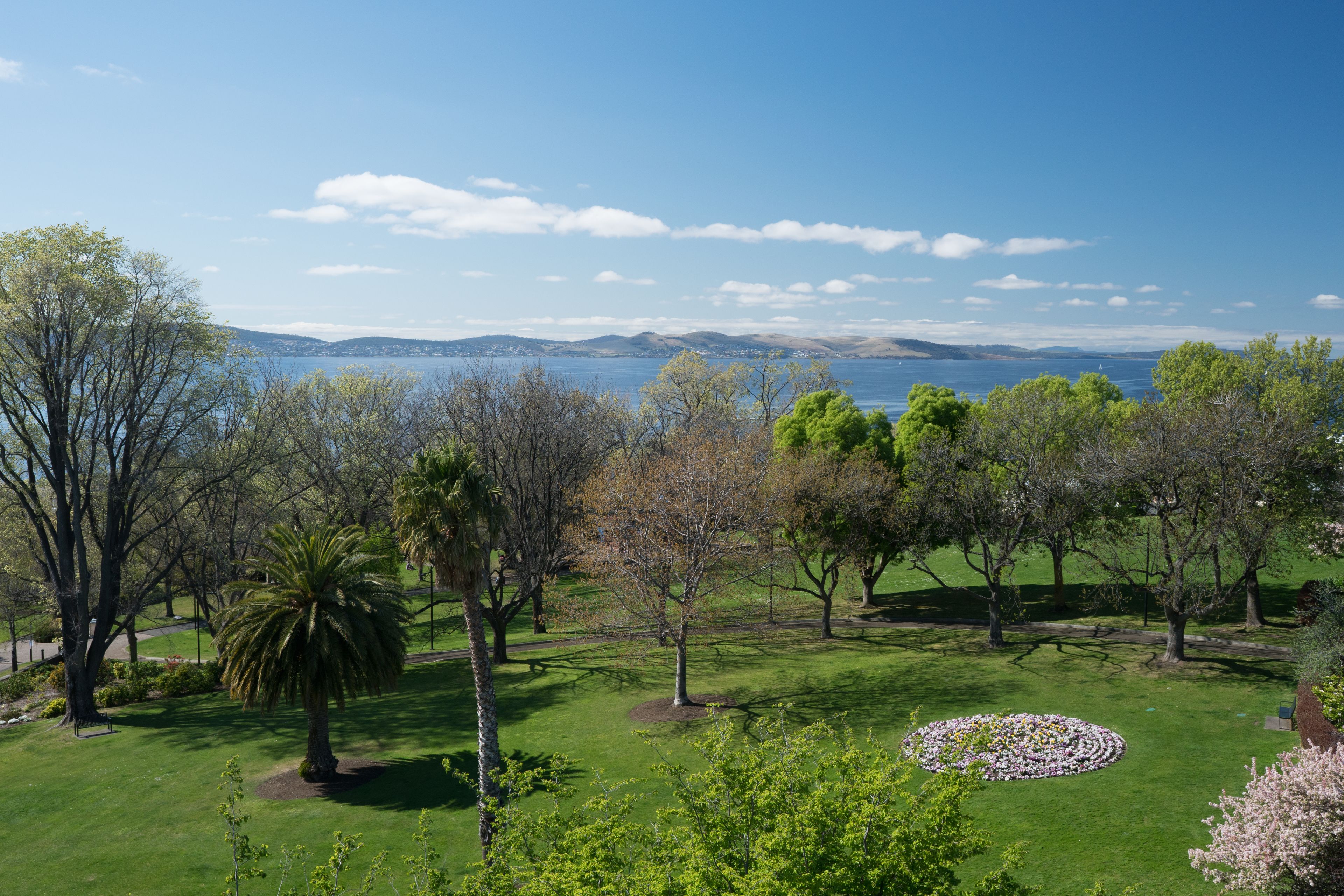 A park with a circular flower arrangement, trees, and a view of the sea.
