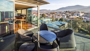 Un balcon doté de meubles en osier et d’une table en verre, offrant une vue sur un paysage urbain avec des montagnes à l’horizon.