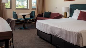 A hotel room with a large bed, a desk, a chair, a small table, and a view of the city through the windows.