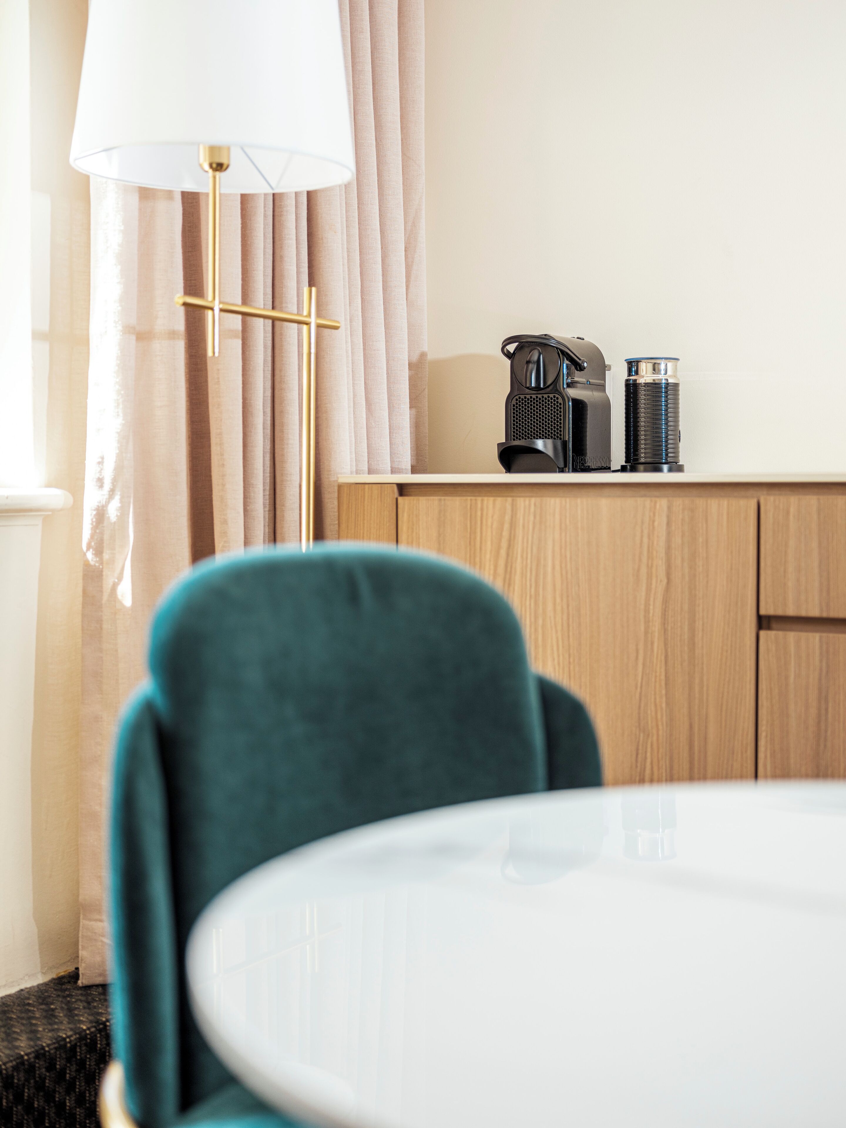 Ett modernt rum med ett runt bord, turkosa stolar, en kaffebryggare och en lampa.