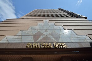 Het exterieur van het Royal Park Hotel met een modern ontwerp, gekenmerkt door een opvallende, ster-vormige glazen gevel en een hoge, cilindervormige structuur aan de rechterkant.