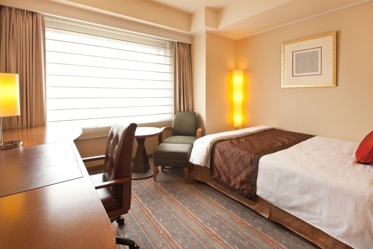 Ein Hotelzimmer mit Bett, Schreibtisch, Stuhl, Lampe und einem Fenster mit Vorhängen.