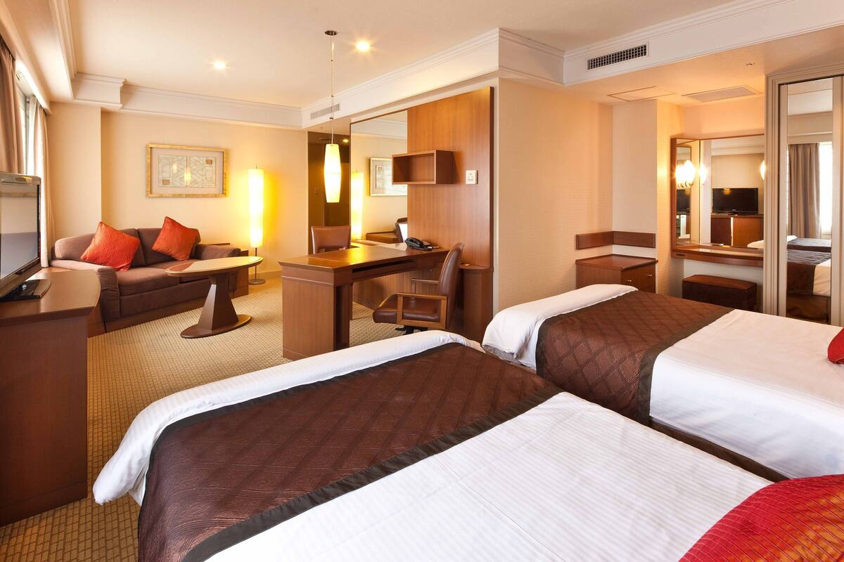 Ein Hotelzimmer mit zwei Betten, einem Schreibtisch, einem Stuhl, einer Couch und einem Fernseher.
