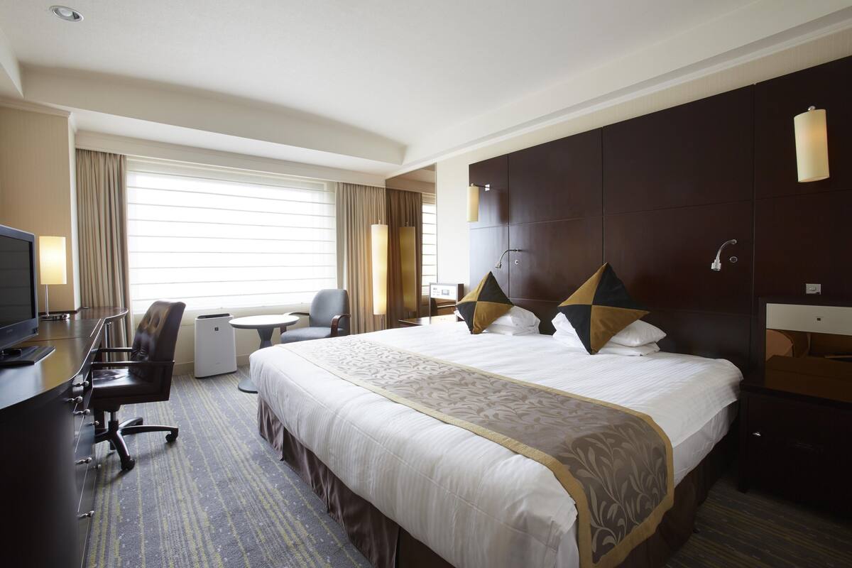 Ein Hotelzimmer mit einem großen Bett, einem Schreibtisch mit Stuhl, einem Fernseher und einem Fenster mit Vorhängen.