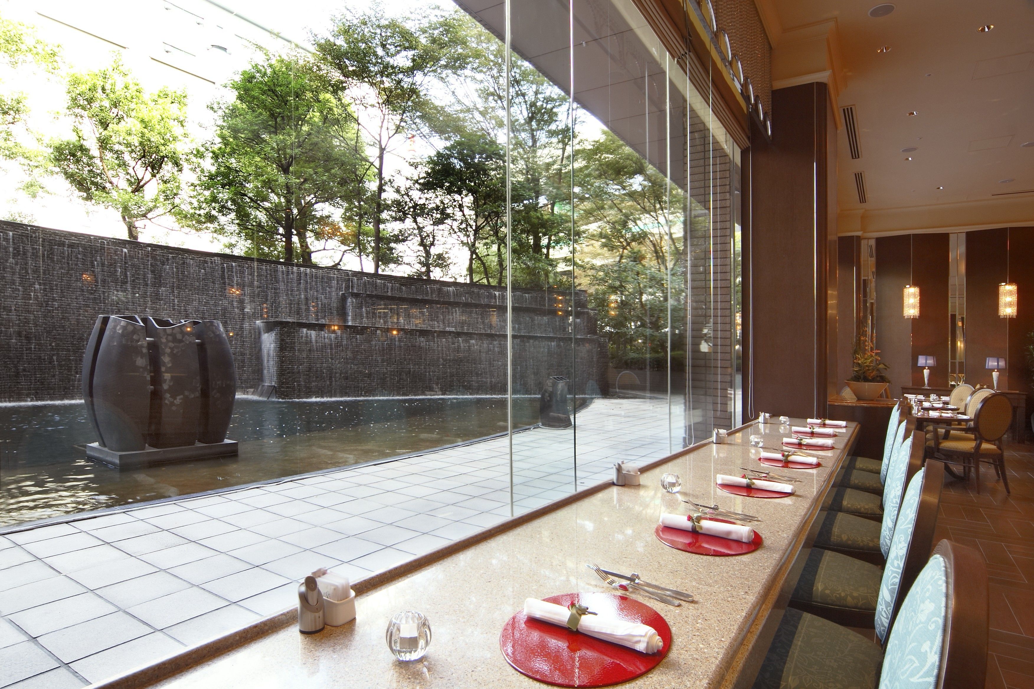 A restaurant with a long table set for dining, a view of a water feature outside, and a brick wall in the background.