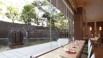 A restaurant with a long table set for dining, a view of a water feature outside, and a brick wall in the background.