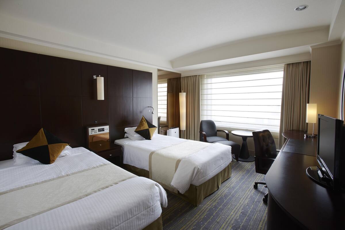 Ein Hotelzimmer mit zwei Betten, einem Schreibtisch, einem Stuhl, einem Fernseher und einem Fenster mit Vorhängen.