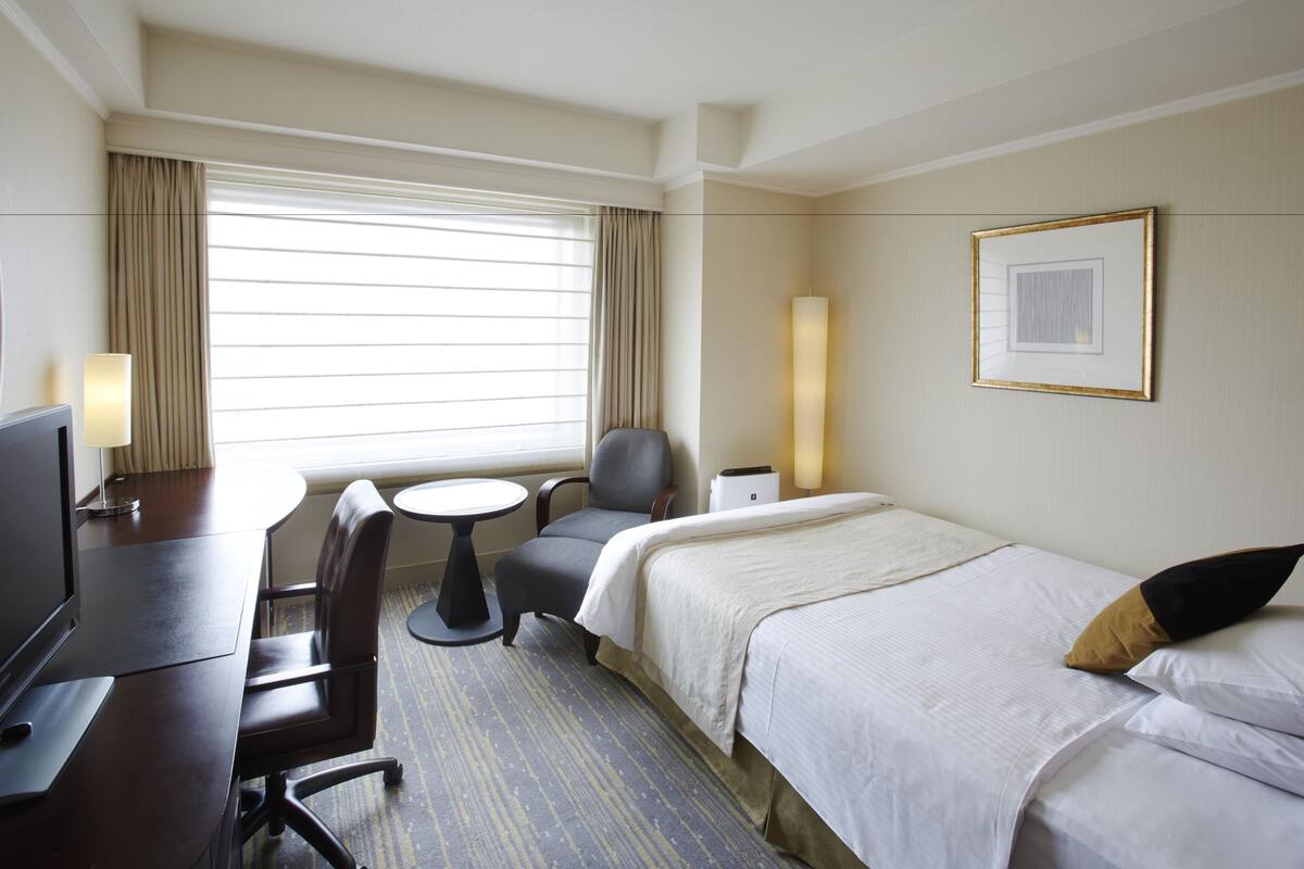 Ein Hotelzimmer mit Bett, Schreibtisch, Stuhl, kleinem Tisch, Lampe und einem Fenster mit Vorhängen.