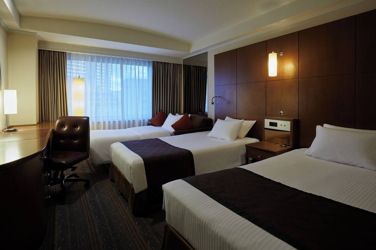 Ein Hotelzimmer mit zwei Betten, einem Schreibtisch, einem Stuhl und einem großen Fenster mit Vorhängen.