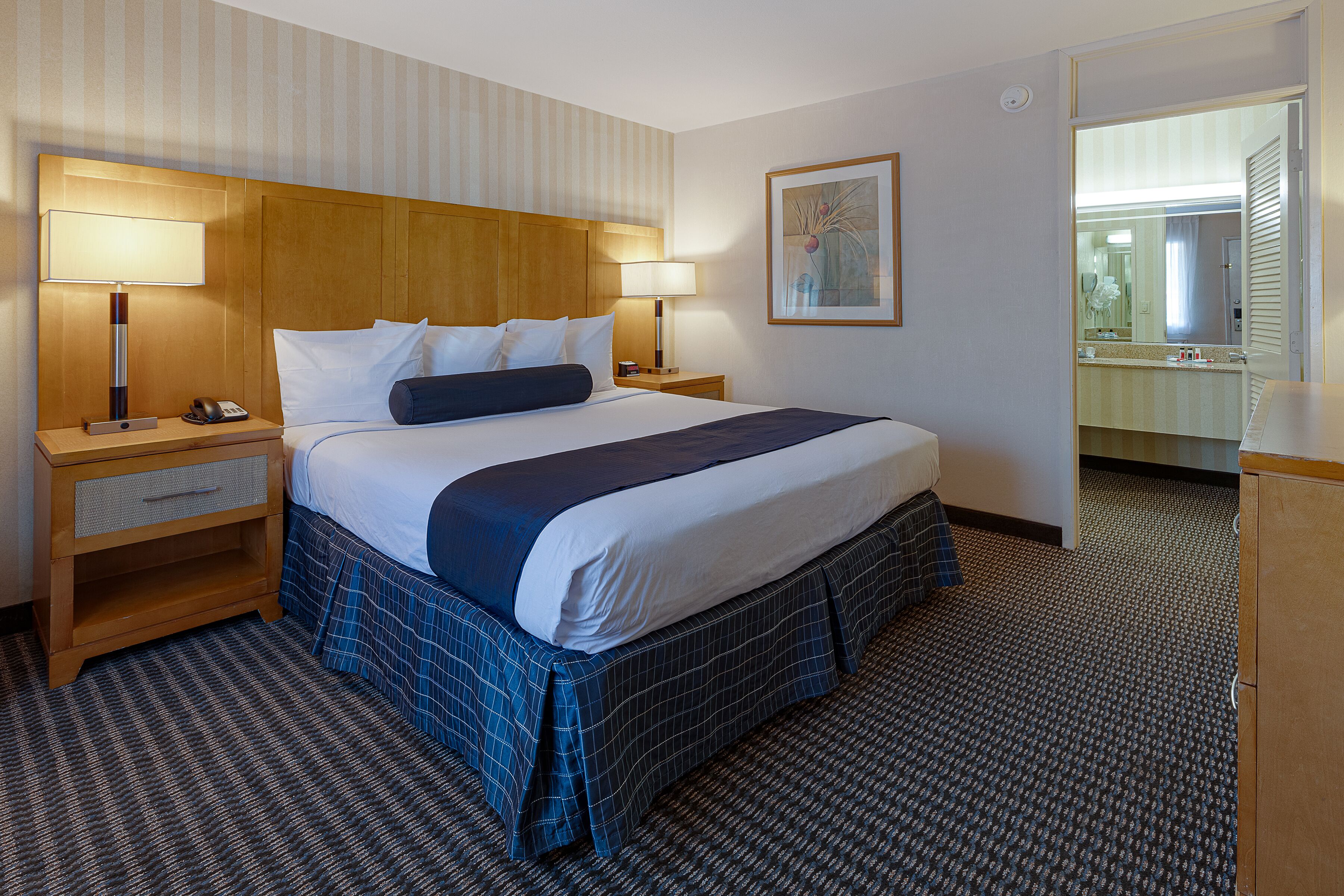 Un cuarto de hotel con una cama grande, dos mesitas de noche con lámparas, un cabecero de madera, una obra de arte enmarcada en la pared, y un baño visible a través de una puerta abierta.