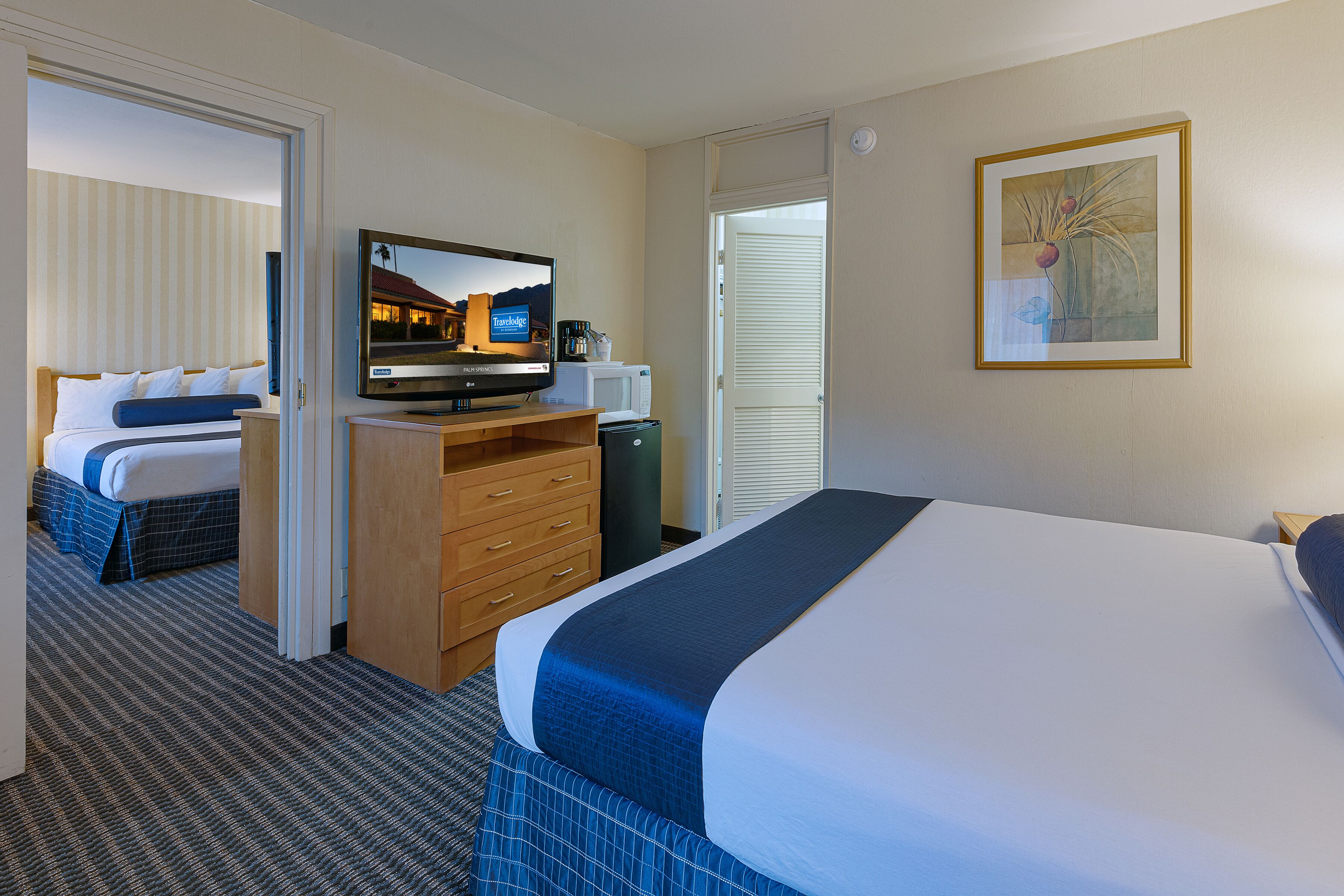A hotel room with a large bed, a television, a desk with a computer, and a door leading to another room.