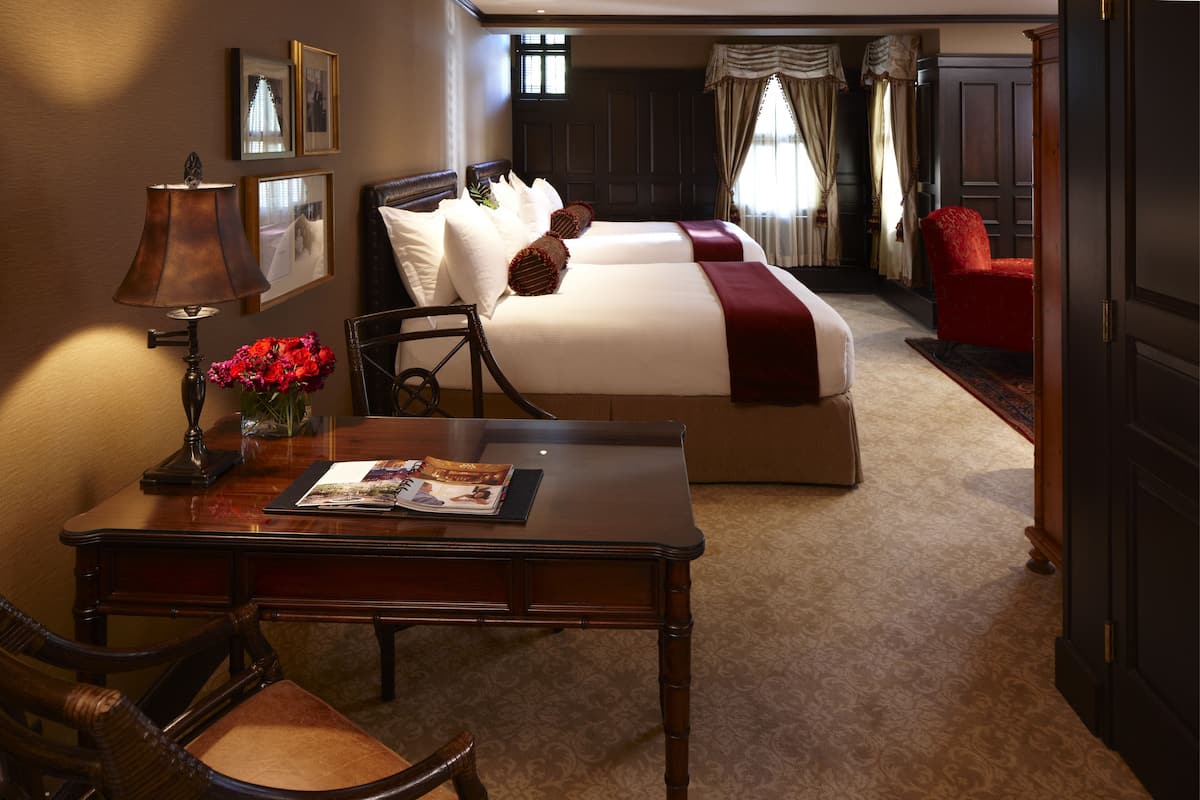 A hotel room with a wooden desk, two beds, a red chair, and a lamp.