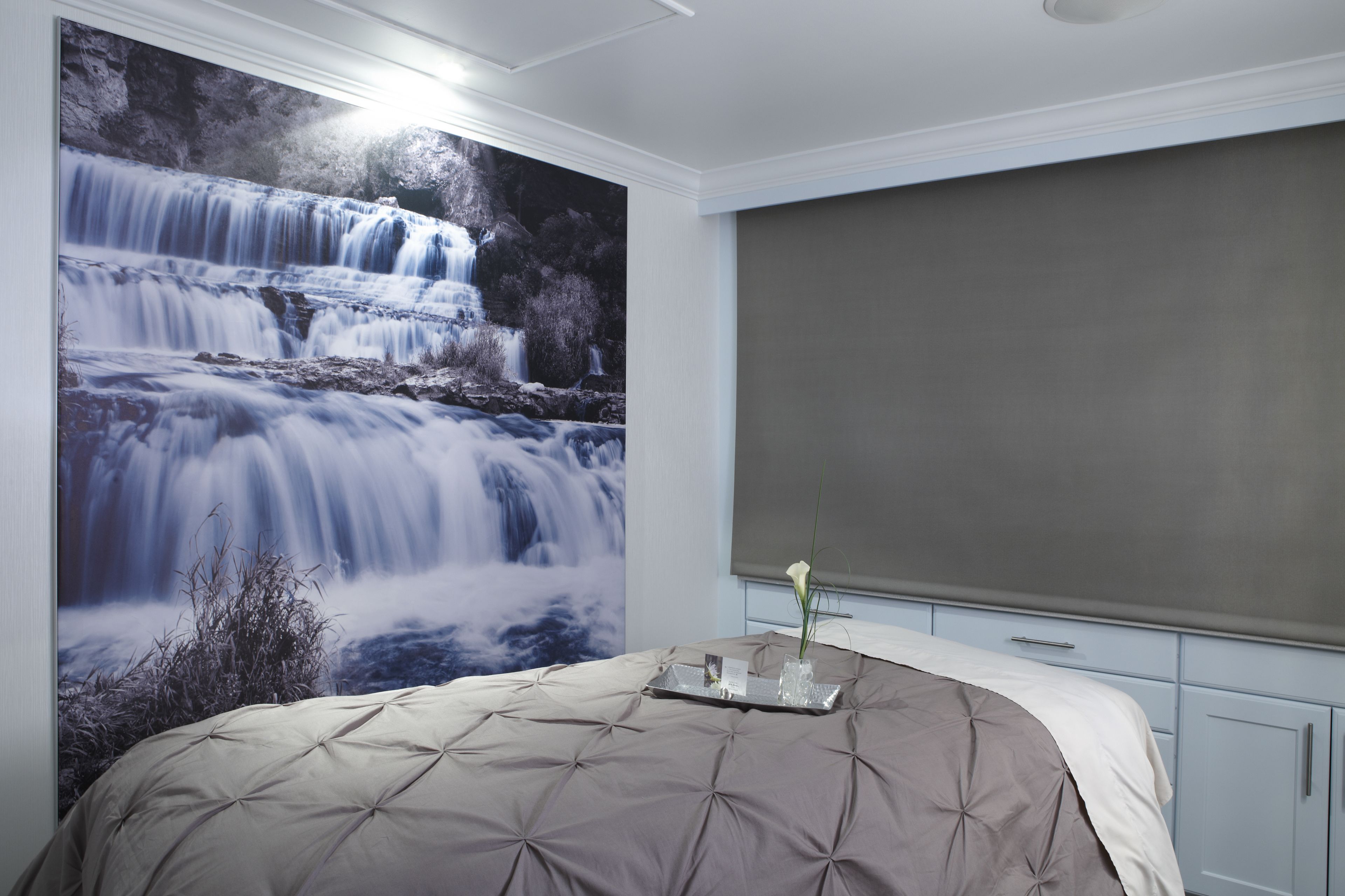 Un dormitorio con un gran mural en la pared de una cascada, una cama con cubrecama gris y blanco, una bandeja con una rosa, y armarios azul claro.