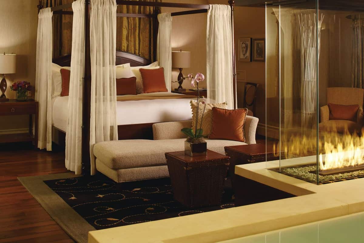 A four-poster canopy bed with a nightstand, a sofa, a glass table with a fire feature, and a chair.