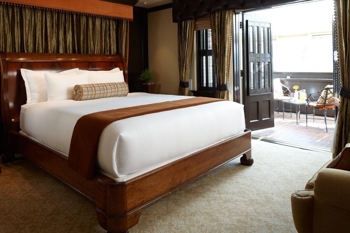 A large bed with a wooden frame and white bedding, a nightstand with a lamp, and a view of the outside balcony.