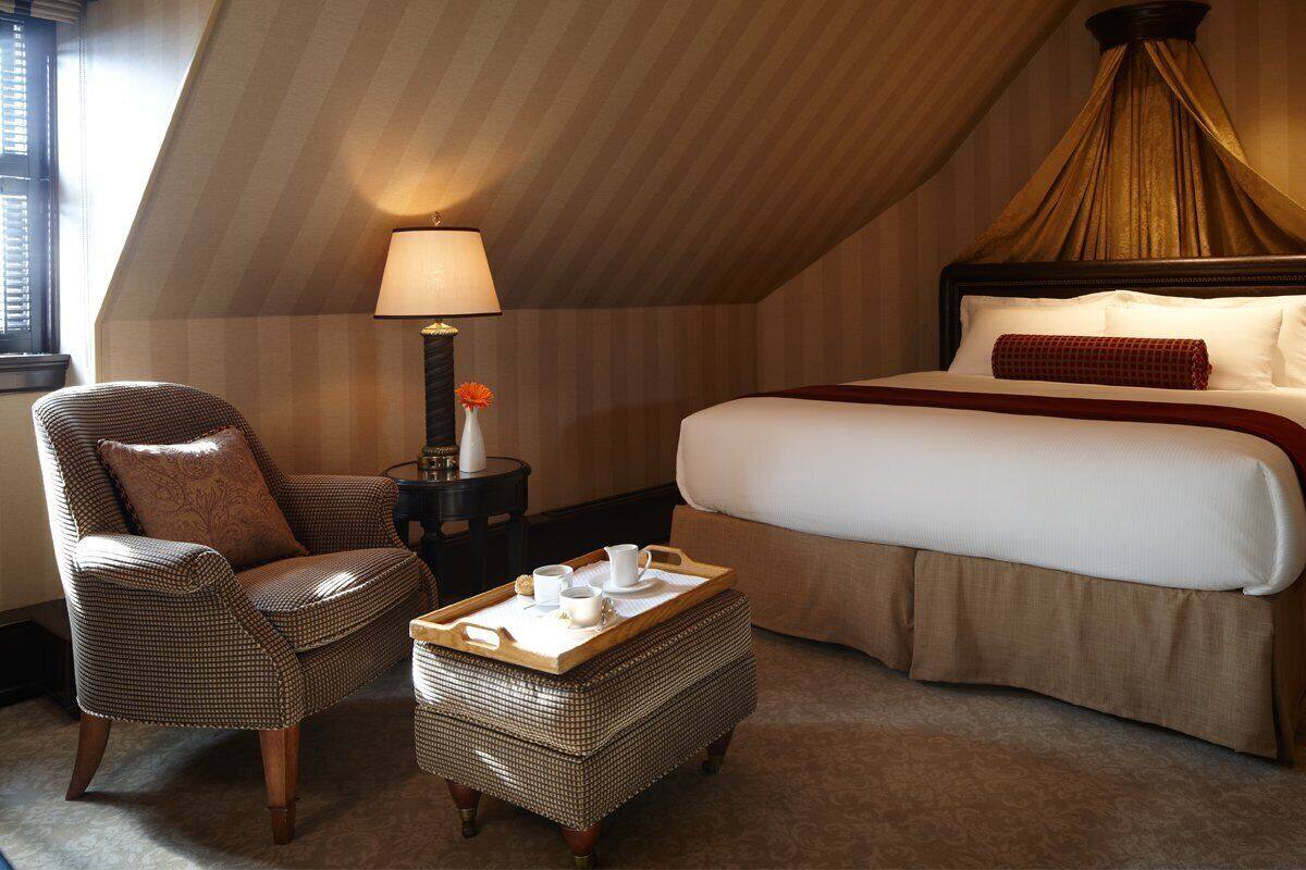 A hotel room with a large bed, a chair, a small table with a tray, and a lamp.