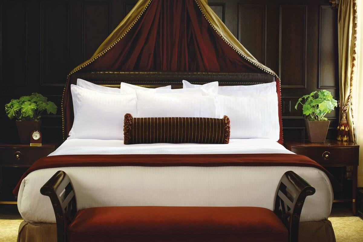 A four-poster bed with white linens and a red canopy, flanked by two nightstands with potted plants.