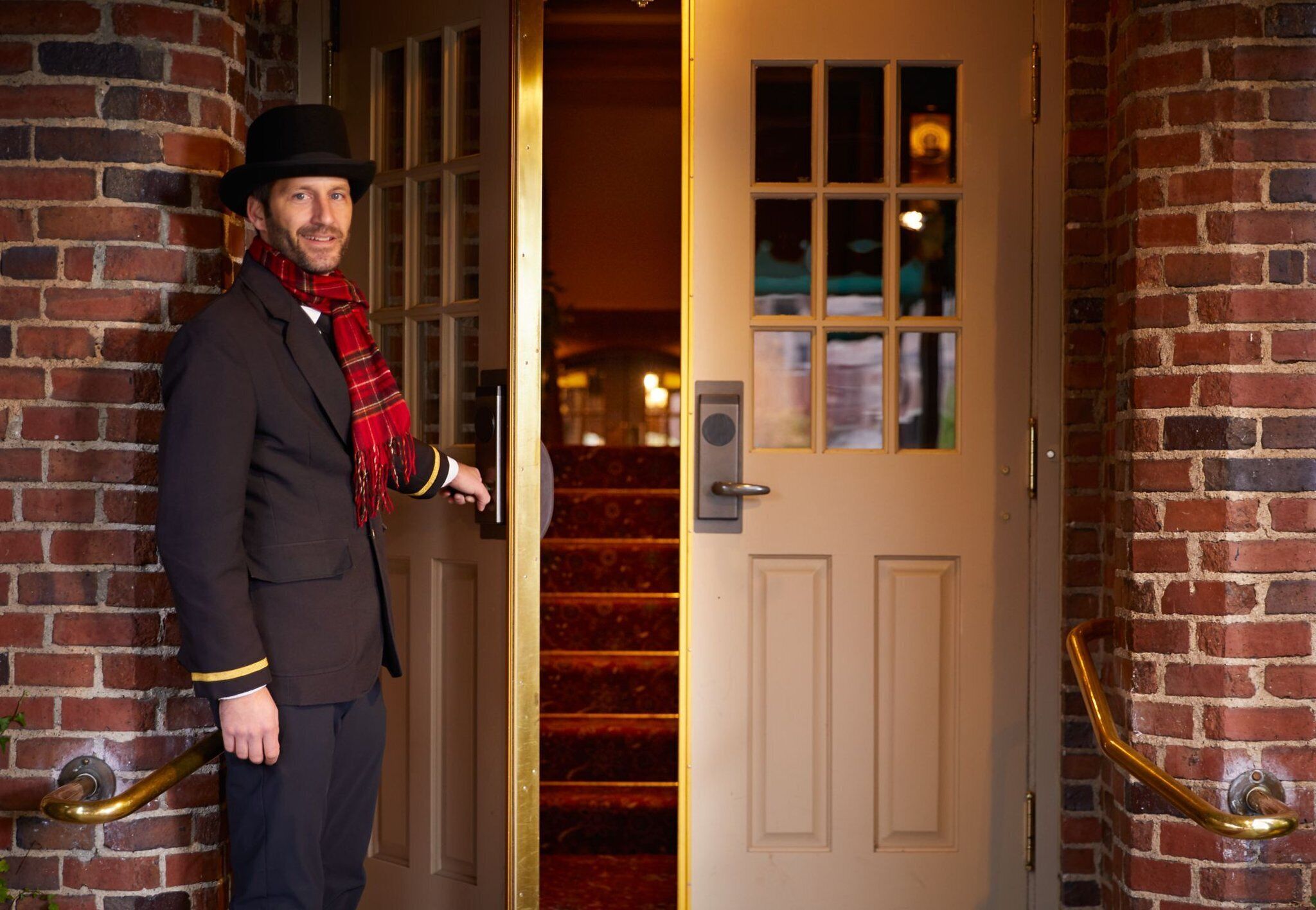 A person standing at a door with a brass handle, a brick wall, and a window with multiple panes.