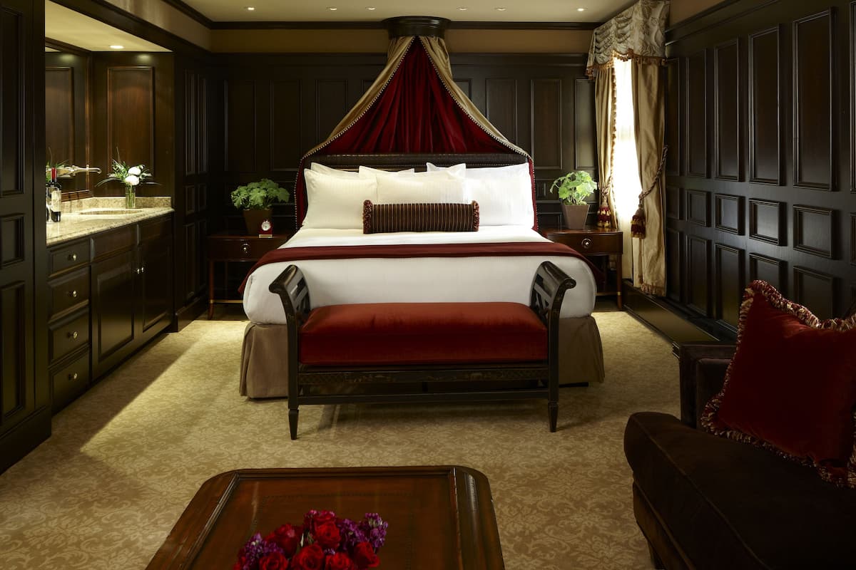 A large, ornate four-poster bed with a red canopy, a dark wood floor, a dark wood sofa, and a small table with flowers.