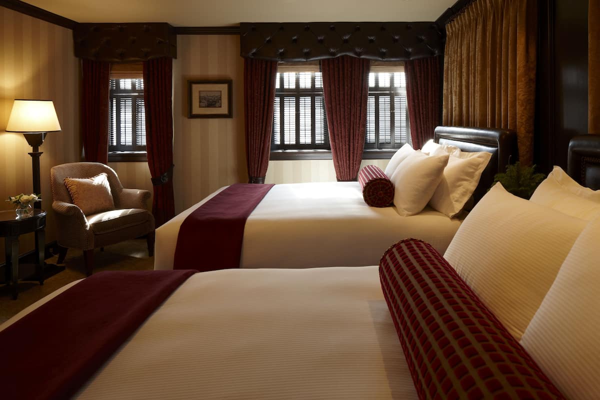 A hotel room with two beds, a chair, a lamp, and a window with curtains.
