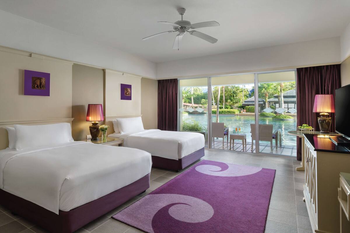 A hotel room with two beds, a ceiling fan, a TV, and a view of a pool and tropical landscape.