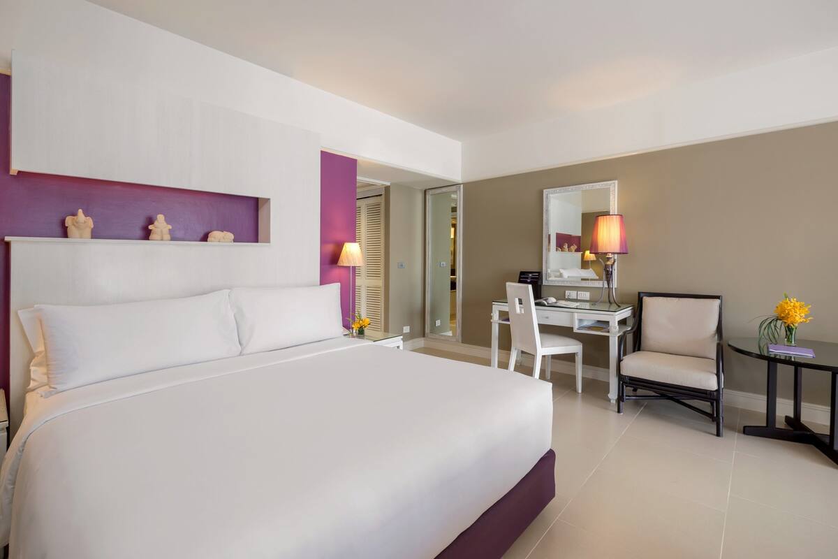 A hotel room with a large bed, a desk, a chair, and a small table with a lamp.