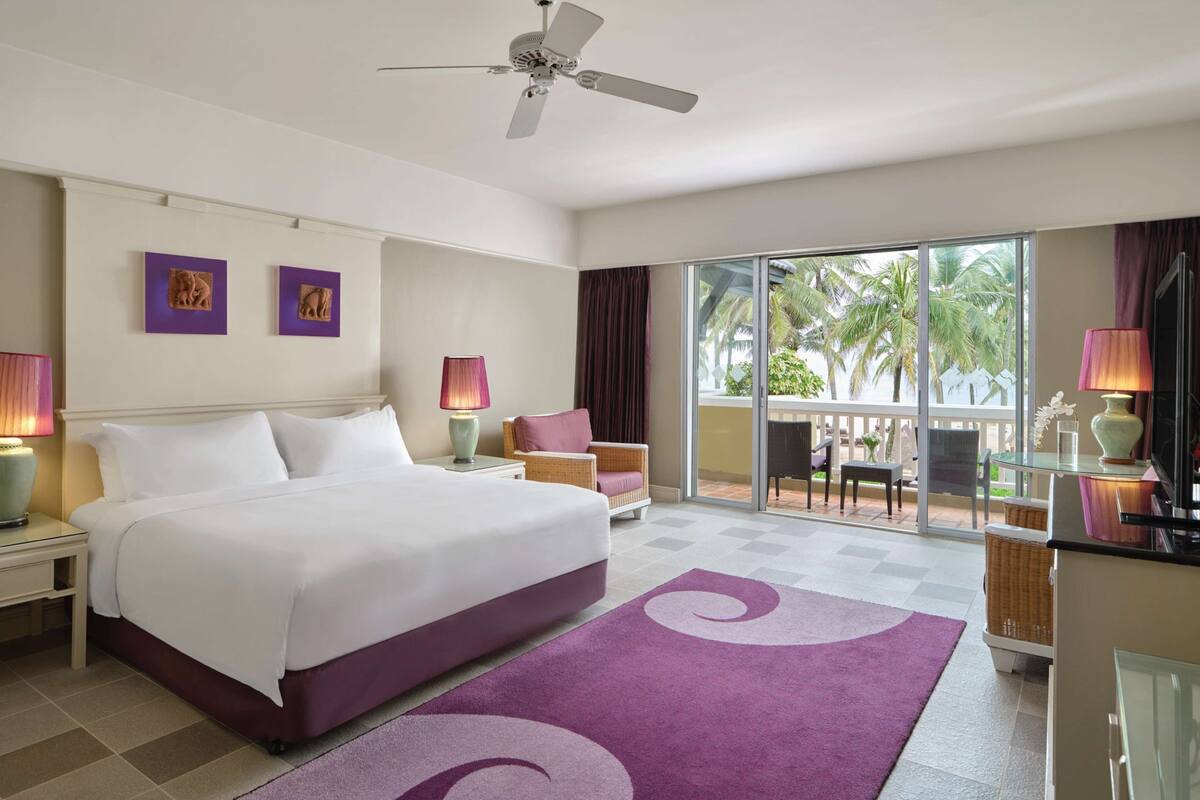 A hotel room with a large bed, a ceiling fan, a balcony with a view of palm trees, and a checkered floor.