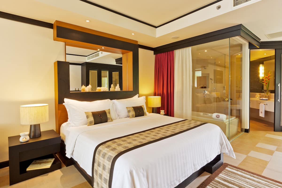 A hotel room with a large bed, a bedside table with a lamp, a glass-enclosed shower area, and a bathroom with a sink and mirror.