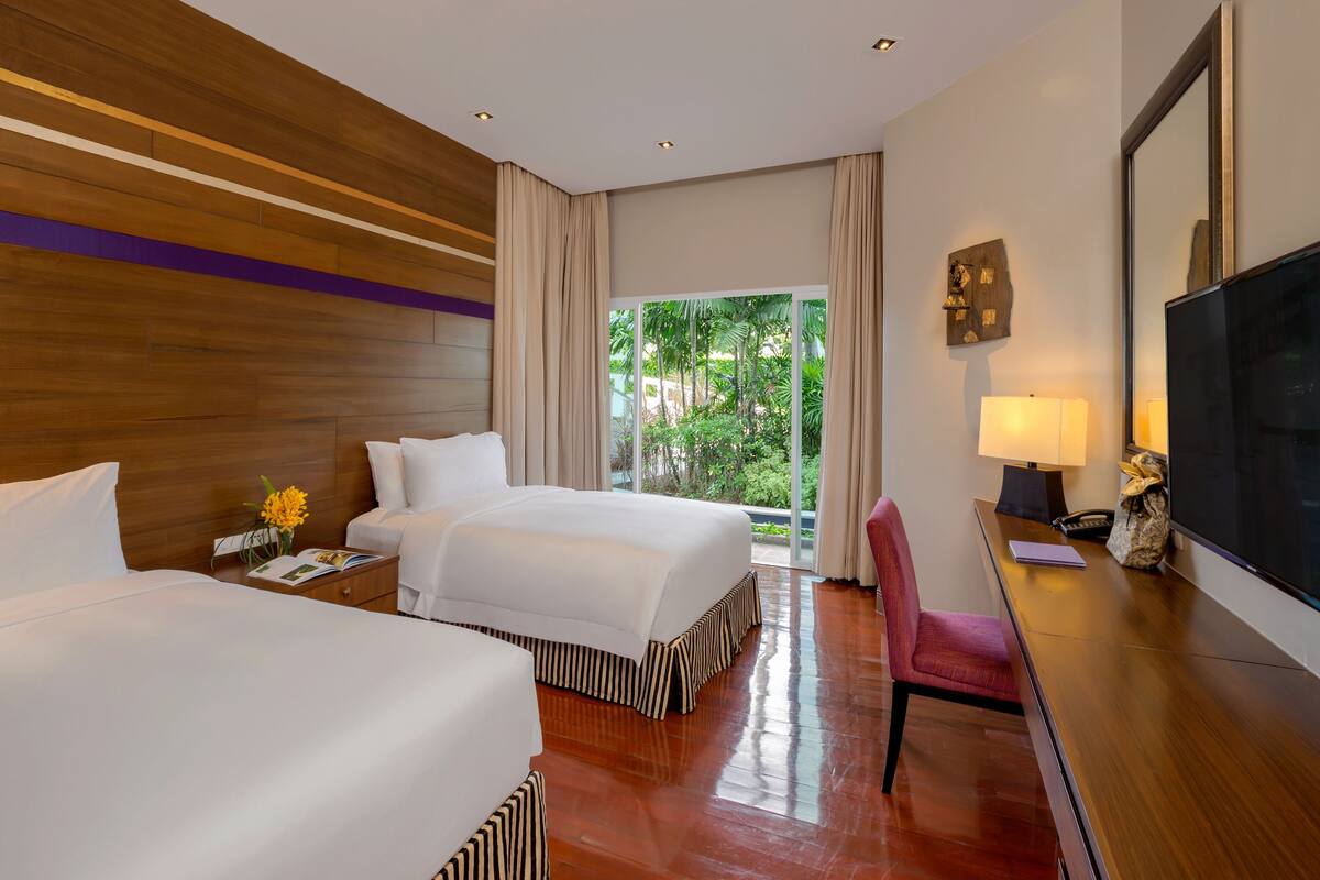 A hotel room with two beds, a desk with a chair, a TV, and a view of greenery through large windows.