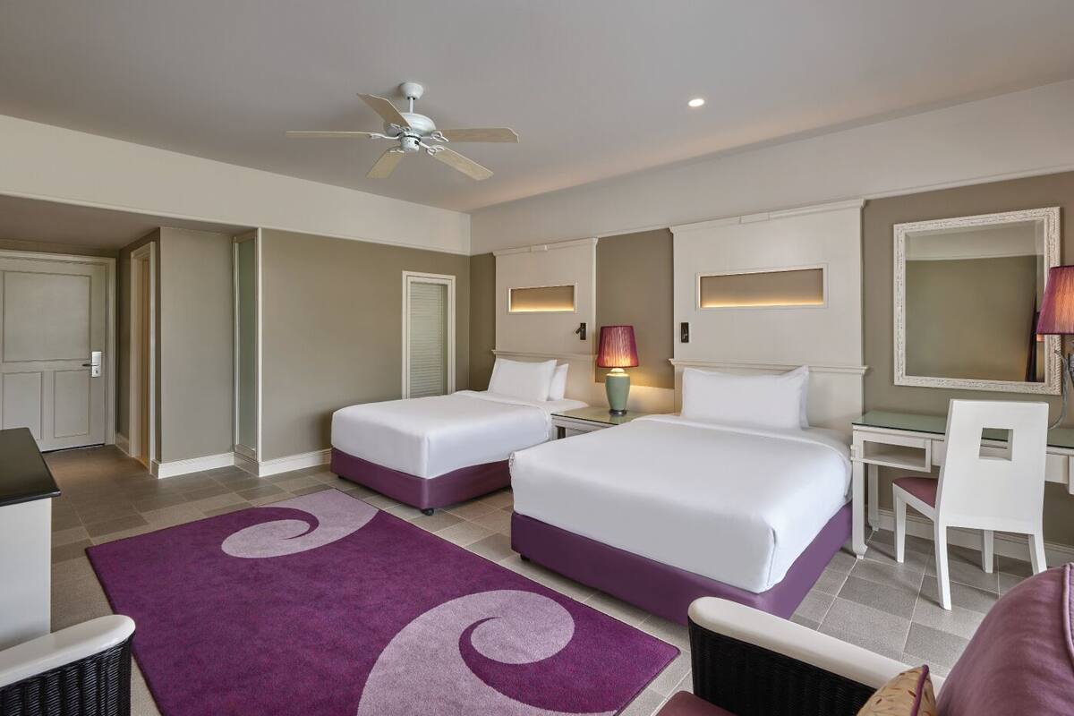 A hotel room with two beds, a desk, a chair, a ceiling fan, and a large mirror.