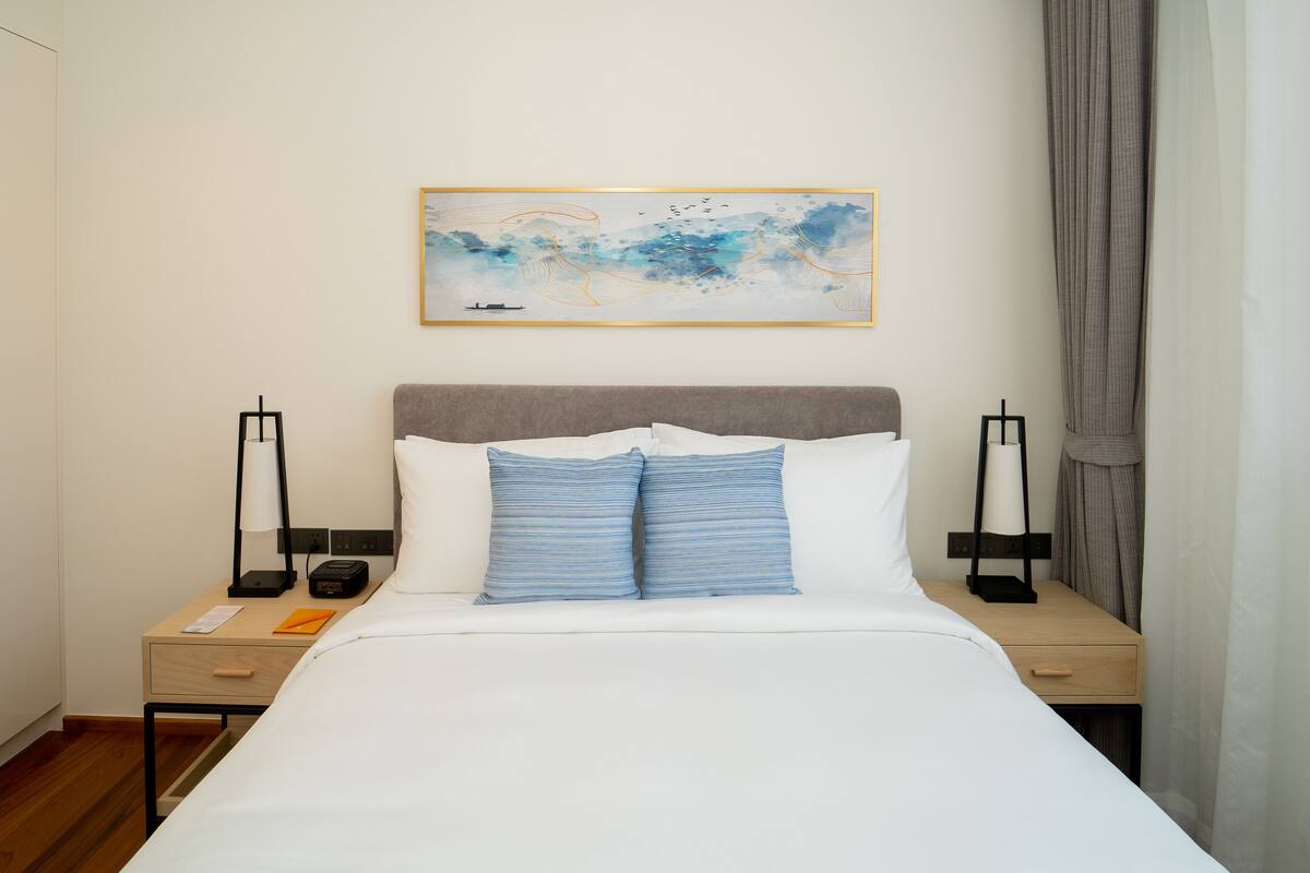 A neatly made bed with a gray headboard, two bedside tables with lamps, a small table with a book, and a framed abstract painting on the wall.