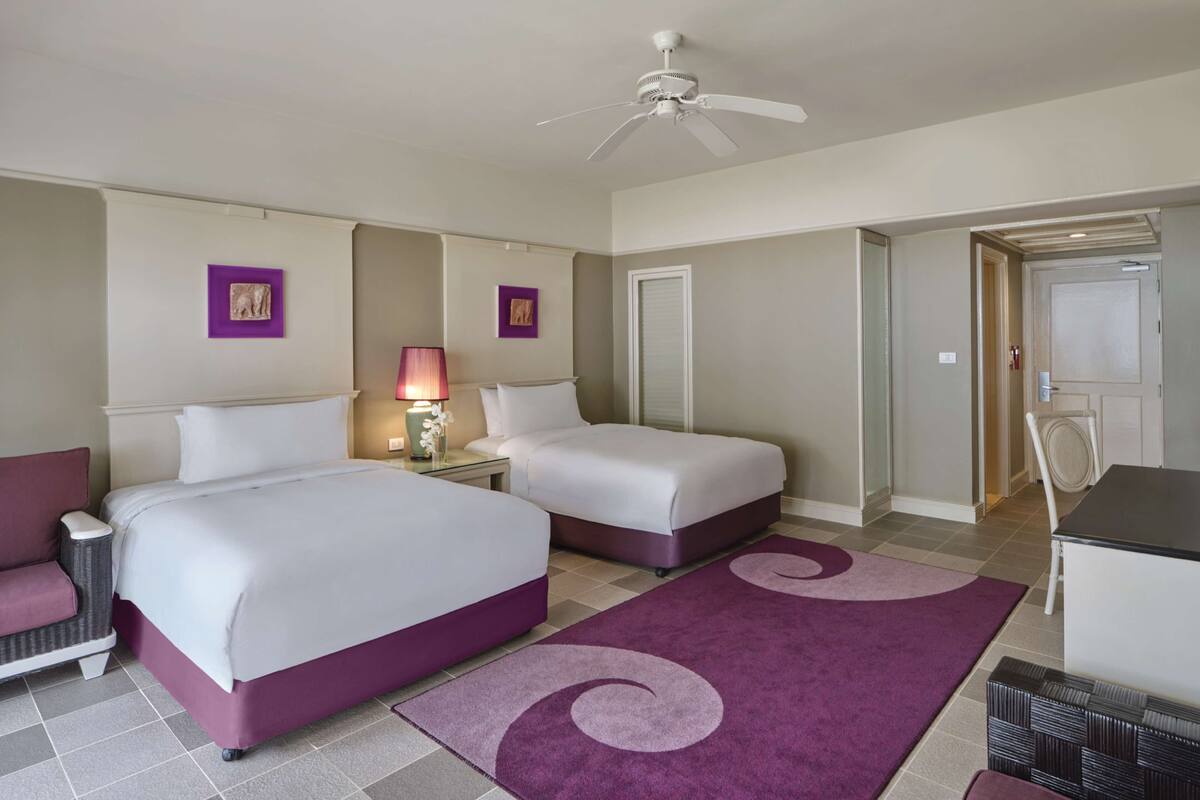 A hotel room with two beds, a desk, a chair, a ceiling fan, and a door.