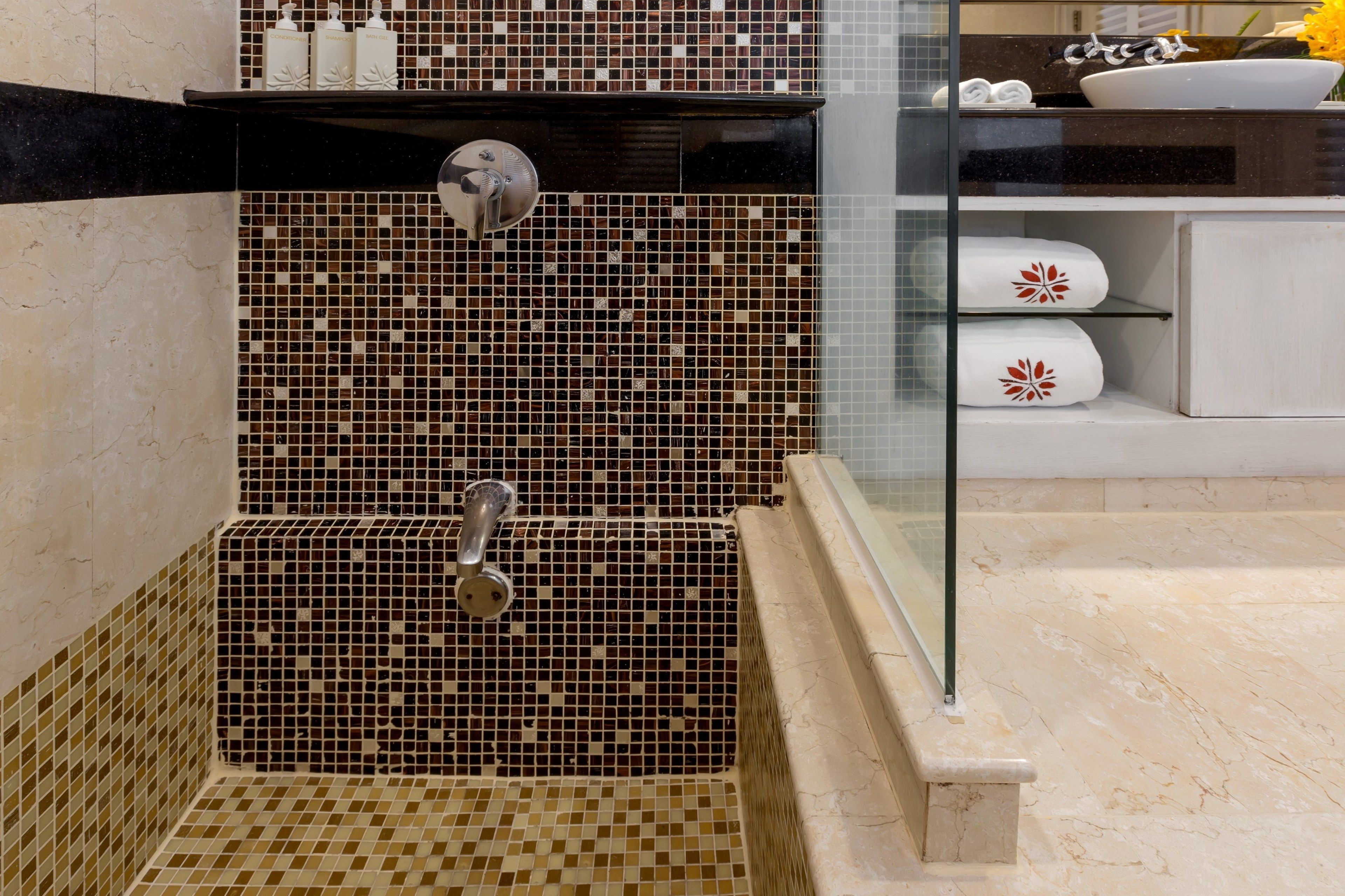 Um banheiro com parede de mosaico, box de vidro e pia branca com espelho em cima.