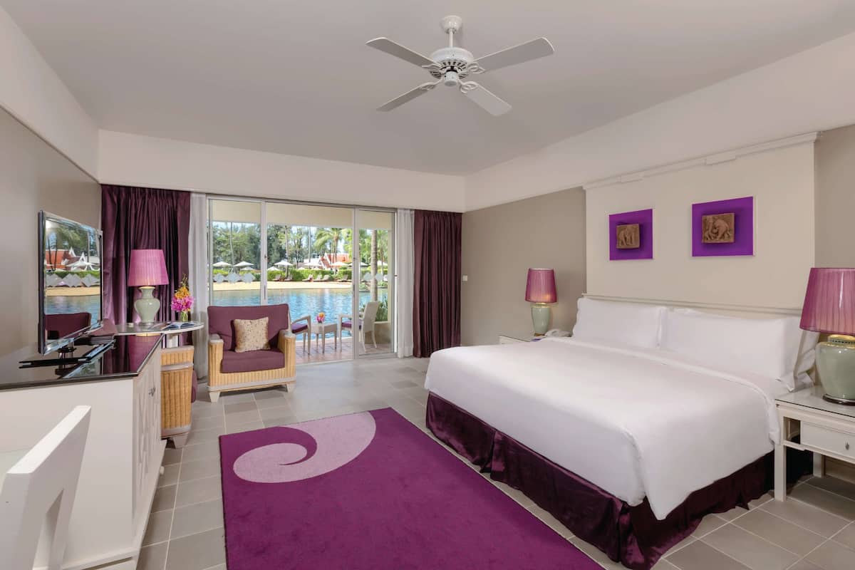 A hotel room with a bed, a desk, a chair, a balcony with a view of the pool, and a ceiling fan.