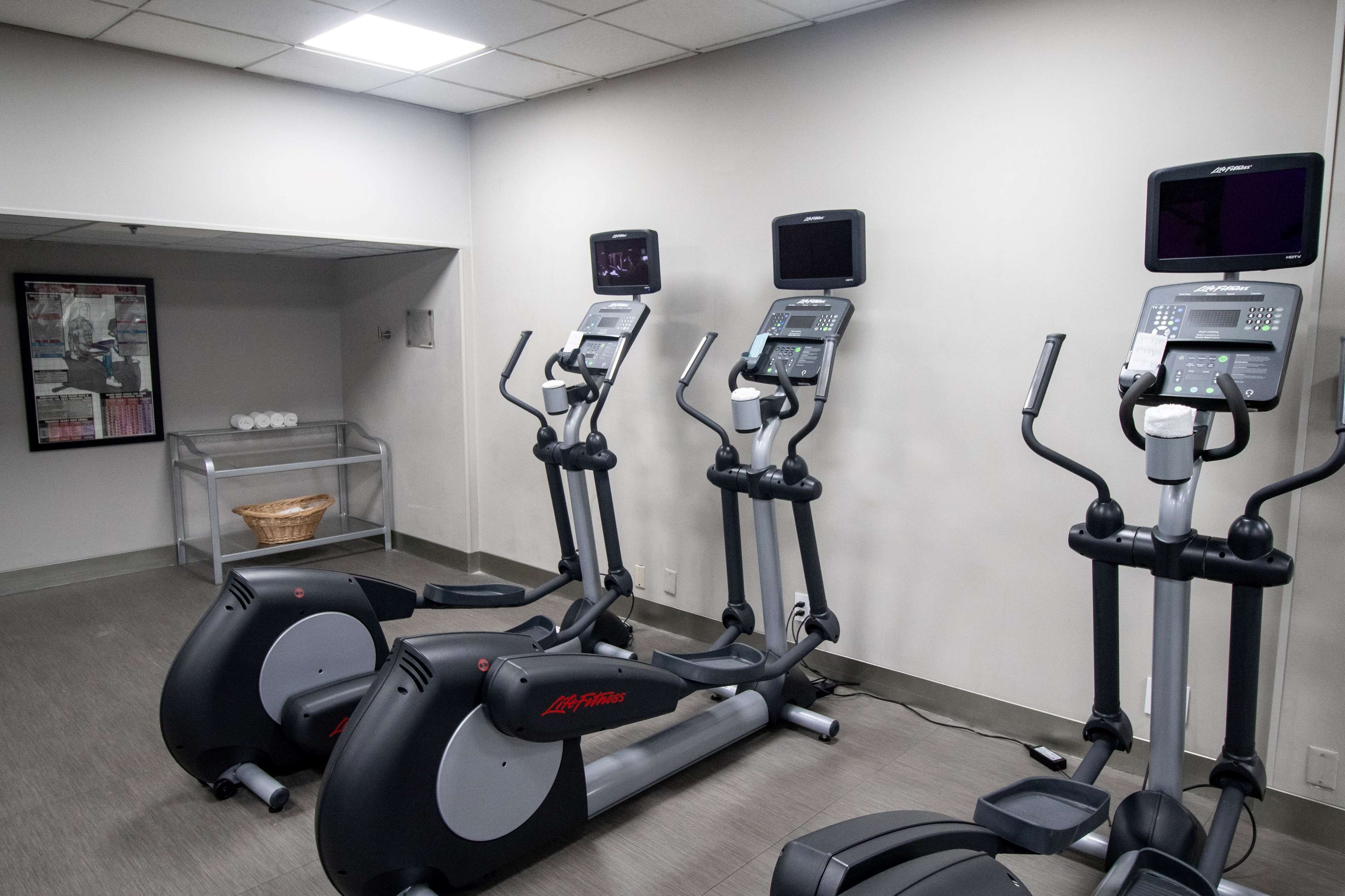 A gym with three elliptical machines, a wall-mounted TV, and a small shelf with a basket.