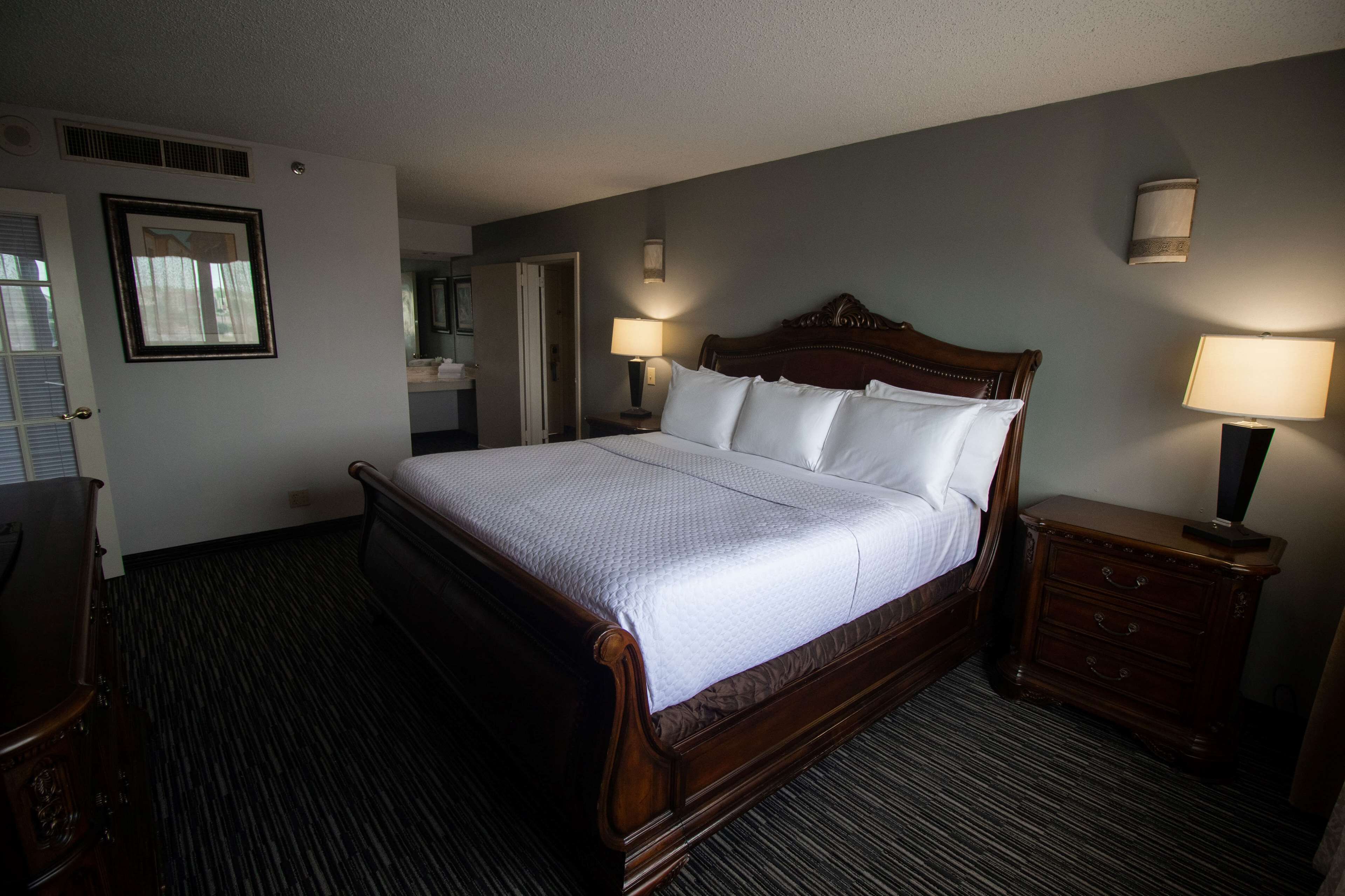 A hotel room with a large bed, two bedside tables with lamps, a dresser, a mirror, and a door.