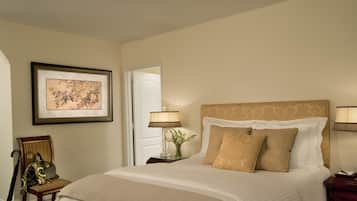 A neatly made bed with a beige bedspread and pillows, a framed floral artwork on the wall, a chair with a handbag, and a lamp on a bedside table.