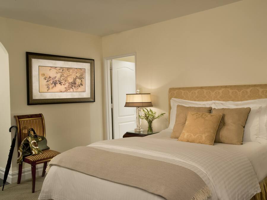 A neatly made bed with a beige bedspread and pillows, a framed floral artwork on the wall, a chair with a handbag, and a lamp on a bedside table.
