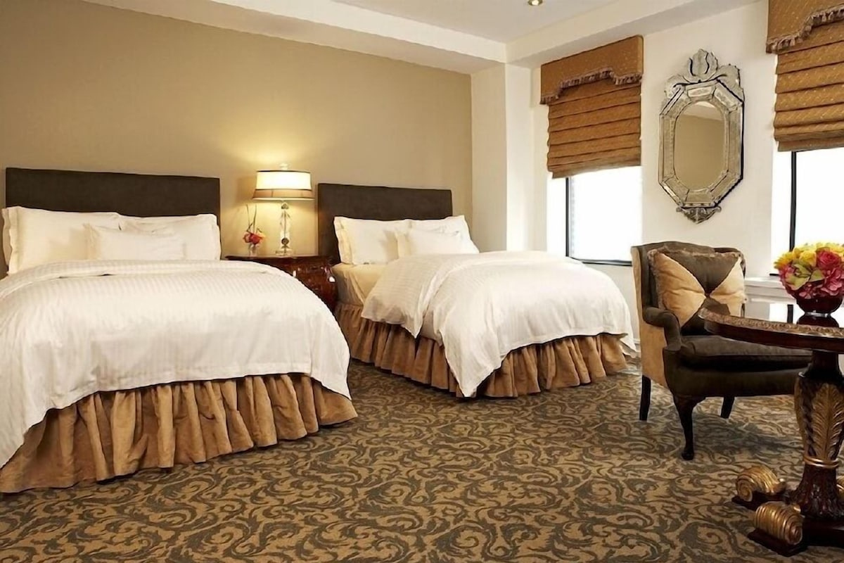 A hotel room with two beds, a chair, a table, a lamp, a mirror, and a patterned carpet.