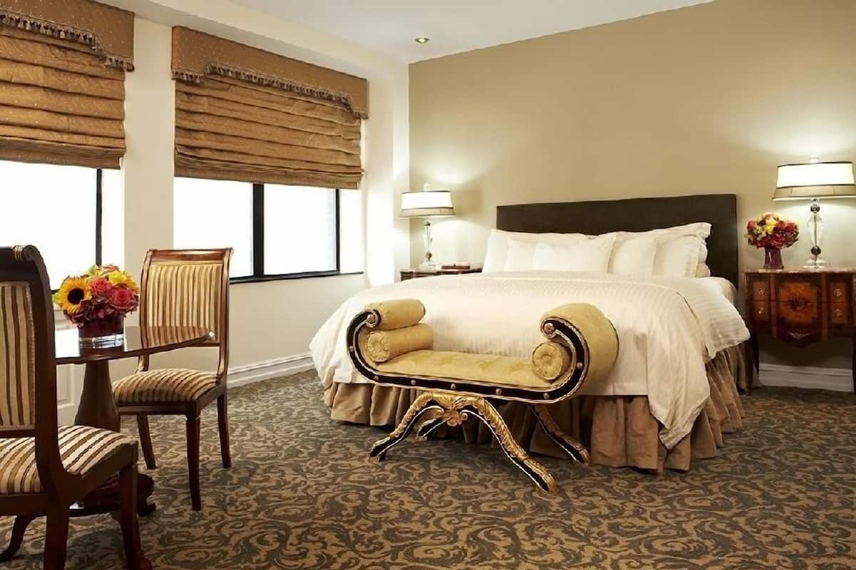 A hotel room with a large bed, a chaise lounge, a wooden chair, a small table with flowers, and a nightstand with a lamp.