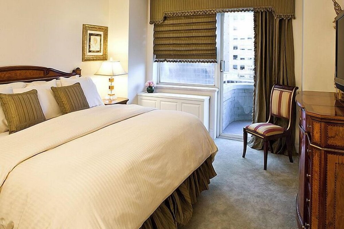 A hotel room with a large bed, a nightstand with a lamp, a chair, a dresser, and a window with curtains.