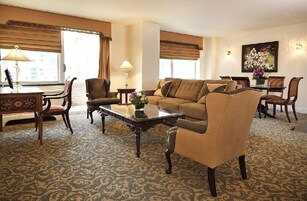 A hotel room with a living area featuring a sofa, armchairs, a coffee table, and a dining table with chairs. There are two lamps, a painting on the wall, and a window with curtains.