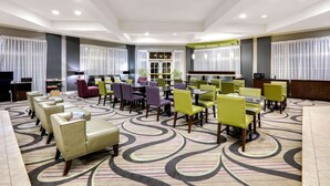 Un hall de hotel con una alfombra estampada, varias zonas de estar con sillas y mesas, y una recepción.