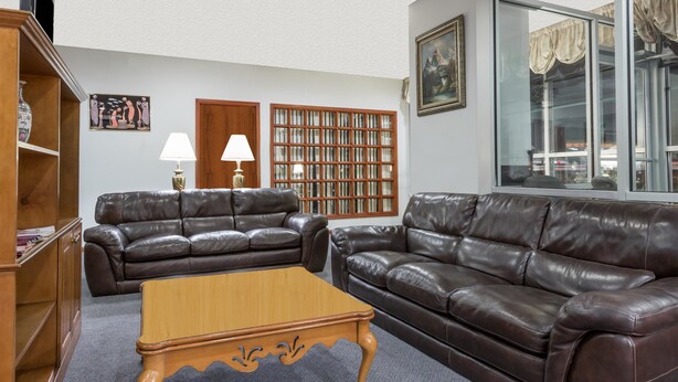 A living room with two brown leather sofas, a wooden coffee table, a bookshelf, a lamp, and a large window with curtains. - Ramada Limited Norfolk (Norfolk)