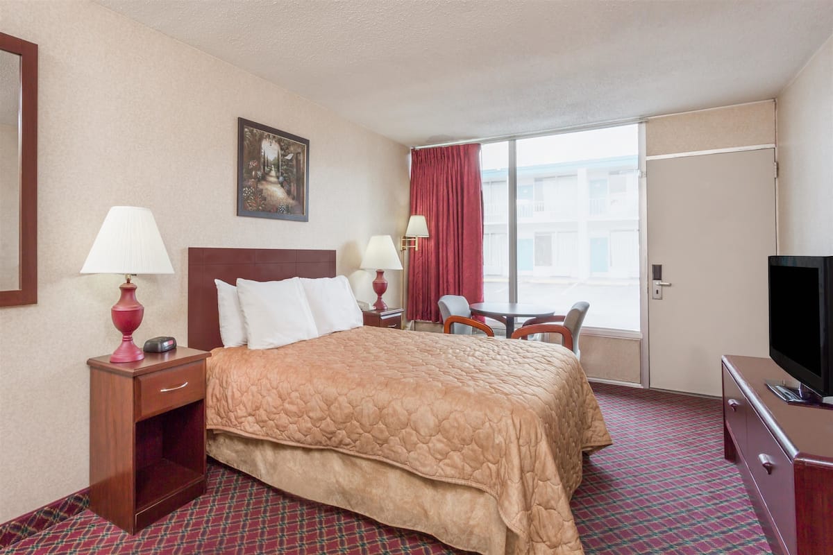A hotel room with a bed, two bedside tables with lamps, a TV, a window with curtains, and a painting on the wall.