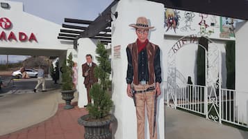 A mural of a cowboy on a building facade, a gated entrance, and a parking area with a car and a person walking.