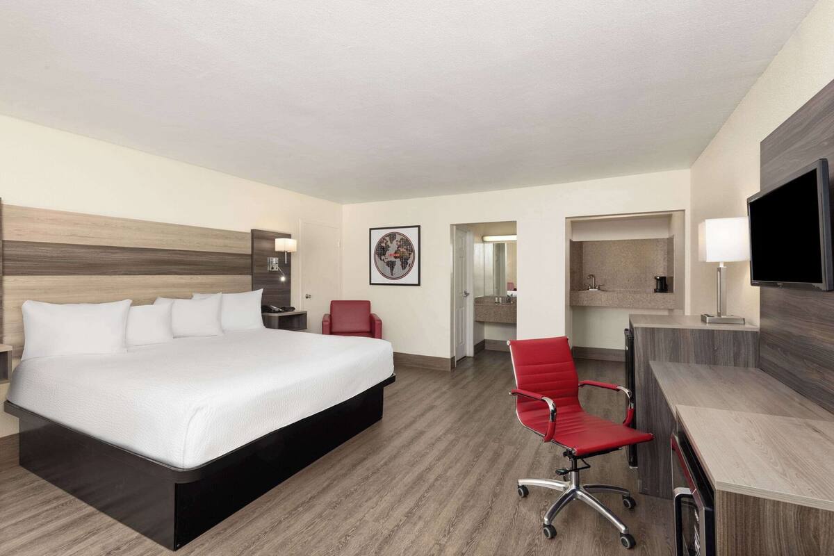 Ein Hotelzimmer mit einem großen Bett, einem Schreibtisch, einem roten Sessel, einem Fernseher und einem durch eine offene Tür einsehbaren Badezimmer.
