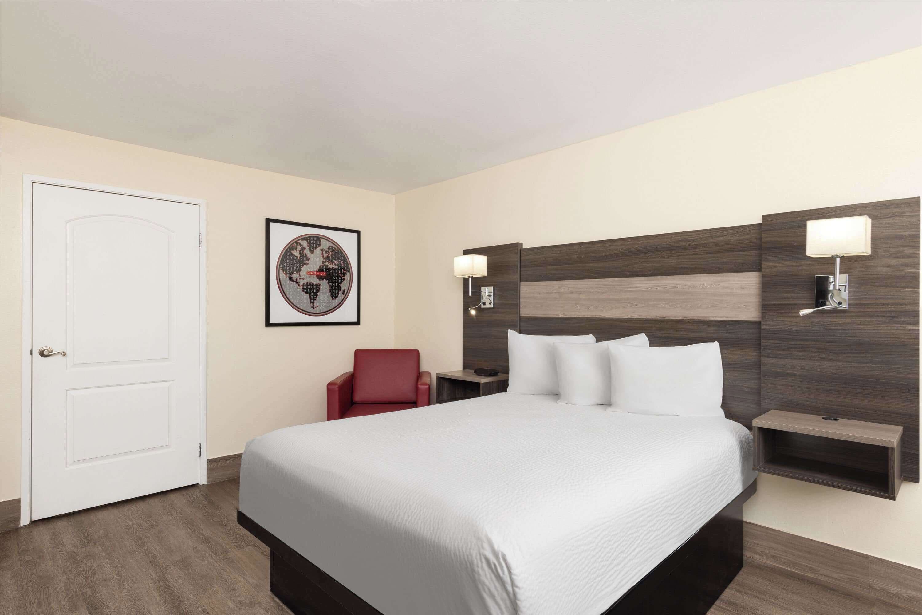 A hotel room with a large bed, a red chair, a framed picture on the wall, and two bedside lamps.